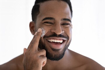 Close up portrait of happy mixed race Black guy applying collagen cream on face, moisturizing,...