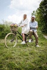 blonde woman with white dress and her boyfriend or husband posing with bicycle with beautifully decorated flower basket in nature and are happy and in love, nature concept, flowers, delivery service