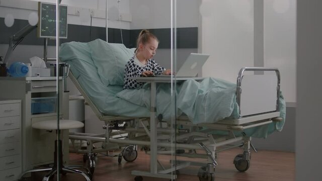 Sick child relaxing in bed playing cartoon video games on laptop computer during medical examination in hospital ward. Hospitalized kid having breathing sickness recovering after surgery