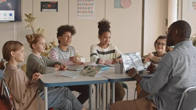 Medium slowmo of cheerful multiethnic schoolchildren and their Afro American teacher sitting in circle at desk in bright modern Science class talking to each other and laughing