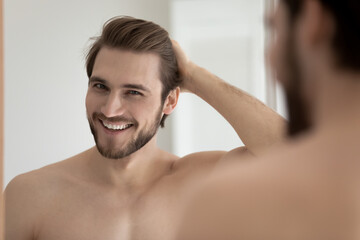 Happy attractive young guy touching smooth silky hair, looking at camera from mirror with toothy smile. Handsome man enjoying haircare, satisfied with shampoo, balm, haircut. Head shot portrait