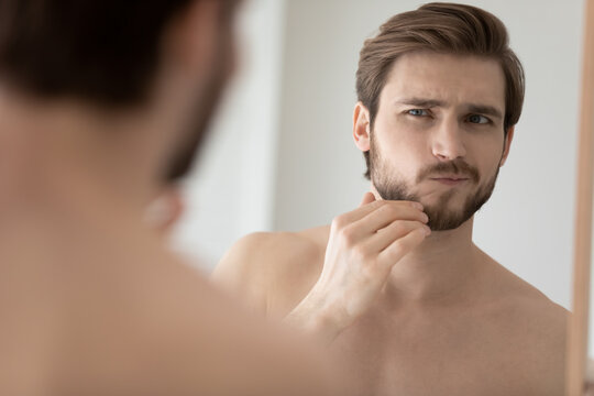 Worry young handsome man examining face in mirror reflection, touching chin and stubble with concerned face, discovering dryness, wrinkles, skin problems, feeling discomfort. Male beauty care concept