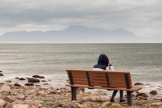 Teenager girl sitting on a bench and enjoys view on Atlantic ocean, low cloudy sky. Croagh Patrick in the background. Travel and tourism concept