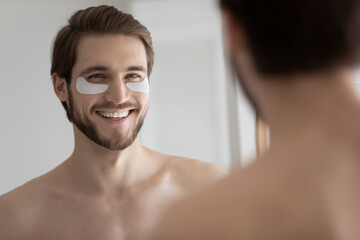 Happy handsome young man applying collagen gel white patches, moisturizing dry under-eye skin, reducing wrinkles, removing bags and circles, refreshing complexion. Male spa, grooming concept