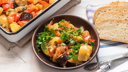 Homemade, rustic vegetarian dish is baked potatoes with eggplant and tomatoes on a plate next to a baking dish.