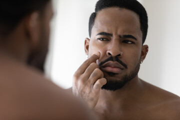 Concerned metrosexual young Black guy with stylish beard, facial piercing worried about face blemish, touching, examining pimple, acne symptom, looking in mirror, Grooming, skincare problems concept