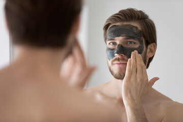 Focused handsome metrosexual guy applying dark cleansing natural clay or mud cosmetic mask on face at mirror for skin treatment, cleaning pores, preventing wrinkles, good complexion. Skincare concept