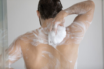 Back of handsome athletic wet young man taking shower, rubbing muscular body with foamy sponge,...