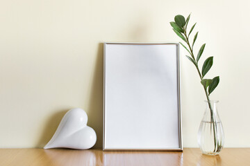 Mockup template with A4 silver frame and white romantic heart, green plant branch in glass vase.