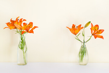 Two vases with orange lily plant flowers in glass vases standing on white surface with blank yellow wall.