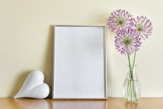 Romantic mockup template with standing A4 silver frame, white porcelain heart and glass vase with purple flowers on wooden shelf.