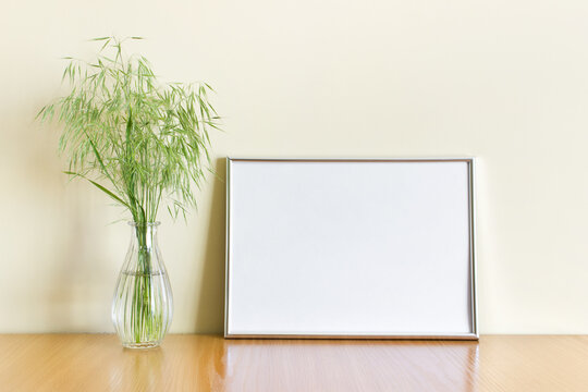 Mockup template with horizontal A4 blank silver metallic frame with green wild forest plants in glass vase on wooden shelf.