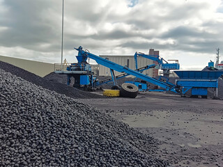 The loading harvester for bulk cargo pours coal onto the bulk carriers at the port quay. Heaps of...