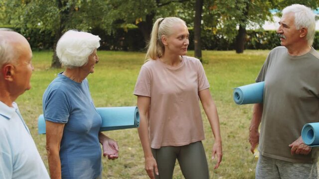 Group of positive senior people in sportswear holding exercise mats and chatting with young female fitness coach while standing together in park