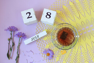 Fototapeta na wymiar Calendar for July 28: cubes with the number 28, the name of the month of July in English , a cup of tea on a yellow openwork napkin, flowers on a pastel background