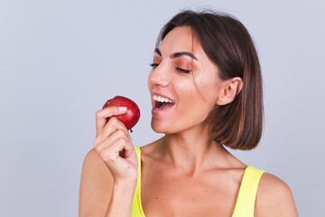 Sports woman stands on gray background, satisfied with the results of fitness training and diet, holding scale, wears top and leggings, holds apple