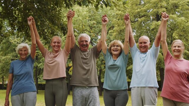 Portrait of young female fitness coach and group of happy senior people in sportswear holding hands, raising arms up and smiling while posing together for camera in park