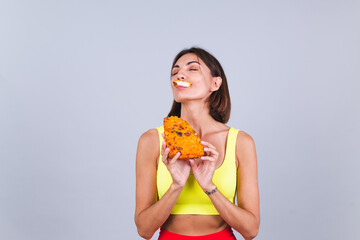 Sports woman stands on gray background, satisfied with the results of fitness training and diet, enjoying  piece of delicious pizza