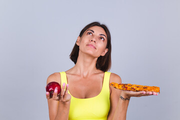 Sports woman stands on gray background, satisfied with the results of fitness training and diet, holds apple and pizza in hands thoughtful