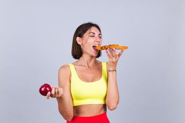 Sports woman stands on gray background, satisfied with the results of fitness training and diet, holds apple and eats pizza with closed eyes enjoy every bite