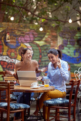 two young adult women sitting in outdoor cafe, smiling, laughing, having online communication with someone