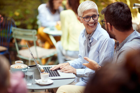 senior  female looking at  young adult beardy man, smiling, with laptop in front of her, while having breakfast in outdoor cafe