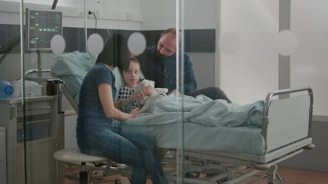 Worried parents sitting with sick girl daughter waiting for sickness expertise during medical examination in hospital ward. Child wearing oxygen nasal tube holding teedy bear while resting in bed
