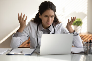 Angry female nurse work on laptop in clinic frustrated by slow internet connection on gadget....