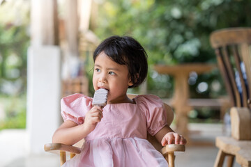 Adorable little Asian girl sitting on wooden chair eating chocolate ice pop