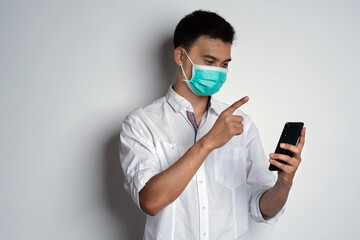 Portrait of young asian man wearing health mask using smartphone with finger pointing to the screen against white background. Getting information in smartphone concept. Novel Coronavirus (COVID-19)