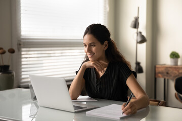 Smiling Caucasian woman sit at desk in office work online on laptop consult client or customer. Happy successful businesswoman busy with computer job at workplace, consider brainstorm with documents.