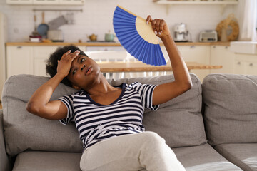 Tired African American woman suffering from heatstroke flat without air-conditioner, waving blue...