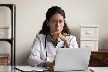 Young Caucasian female doctor in white medical uniform look at laptop screen work online in modern private hospital. Pensive woman GP use computer in clinic consult patient distant on gadget thinking.