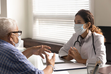Young Caucasian female doctor in facial mask and old male patient talk discuss disease anamnesis in hospital. Woman GP and mature man in facemasks have consultation in clinic in covid-19 corona times. - obrazy, fototapety, plakaty