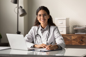 Portrait of smiling young Caucasian female doctor or nurse in white medical uniform in clinic fill journal. Happy woman GP work online consult patient on computer in hospital. Healthcare concept.