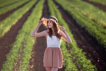 Young woman in white T-shirt, powdery shorts runs along green plowed field. Long hair develops at sunset, the girl has straw hat. Promote agricultural perspective, the texture of black soil ground