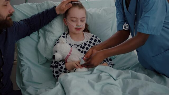 Worried father sitting with hospitalized sick daughter during medical examination in hospital ward. Specialist pediatrician doctor monitoring temperature to hospitalized child with breathing disease