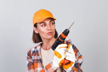 Beautiful woman in plaid shirt, hat and gloves, on gray background holds drill to repair  home confident happy smile