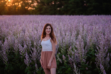 Young woman in white T-shirt, powdery shorts stands by blooming pink sage field. Portrait girls in the orange rays of the sun, hair fluttering in the wind at sunset. Flower texture. Travel out of town