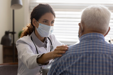 Caring female GP in facial mask touch support old male patient hear bad negative diagnosis at consultation. Woman doctor in facemask comfort caress mature man in hospital, show good medical service.