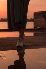 a girl in a green dress walks through puddles after rain at sunset near the river. Girl's legs in snickers. Selective and soft focus