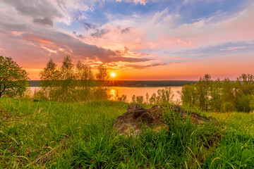 Scenic view at beautiful sunset on a shiny lake with old rough stone on the foreground, green grass, birch trees, golden sun rays, calm water ,nice cloudy sky on a background, spring landscape