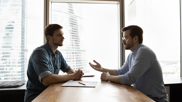Concentrated young male vacancy candidate asking questions about job position to confident employer at interview meeting. Professional serious financial advisor giving consultation to client.