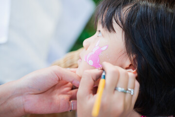 Asian girl sitting for painting face during Easter. An adult's hand is drawing a rabbit cartoon on a cute child face. Children 4-5 years old are engaged in Easter activities.