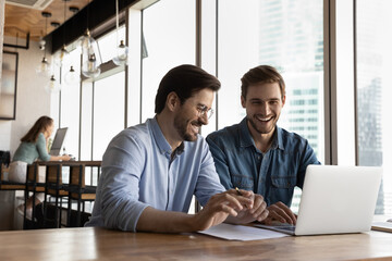 Happy sincere friendly young male colleagues laughing, holding pleasant conversation or watching...