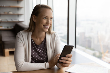 Happy attractive young business lady manager worker dreaming looking in distance, getting message with pleasant news on smartphone, thinking of new challenges or future opportunities in office.