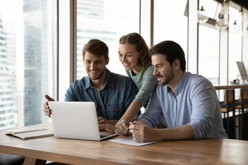 Happy three teammates working together on online project, sitting at table in office room. Smiling...