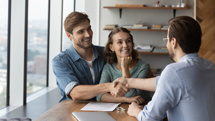 Happy loving married family couple shaking hands with professional real estate agent or broker, thanking for professional consultation, making deal, celebrating purchasing own dwelling apartment.