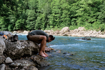 Young handsome Caucasian male traveler with dreadlocks and beard washes his feet in clean cold mountain river and sits on rock. Getting legs wet in icy river. Man is tourist.