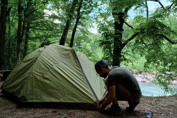 Put tourist tent in campsite and prepare for rest. Young handsome Caucasian male traveler with bandana on his head and beard sets up tent for rest in clearing in forest. © Ekaterina